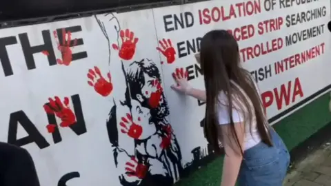 A young woman in a white top and blue jeans places a red hand print on a gable wall
