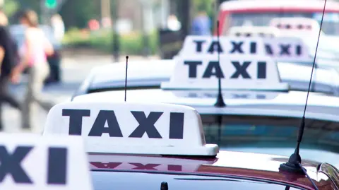 Row of taxis lined up on a city street, each with a roof sign displaying the word ‘TAXI