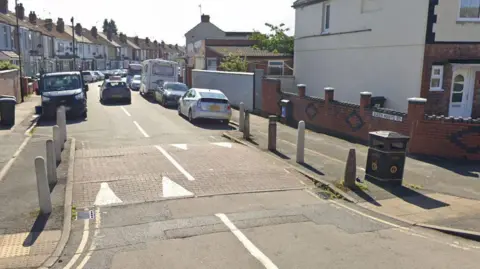 Google A Google street view image of Queen Mary's Road. The road sign is on a low red brick wall to the right and rows of terraced houses can be seen on both sides of the street, with cars parked outside them on the road.