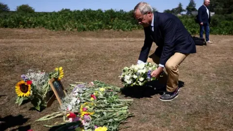 Reuters Nigel Farage, wearing a navy blazer, blue shirt, beige trousers and boat shoes is seen bending down to place a wreath of cream and lilac flowers. On the ground, bunches of flowers have already been laid, alongside a framed black and white photograph of Ann Widdecombe