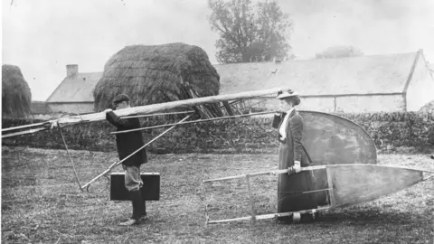 Philip Jarrett A black and white image of Percy and Ella Pilcher, both in Victorian-style dress, carrying the dismantled Bat glider through a grass area. A house or barn building can be seen in the background behind a small stone wall.