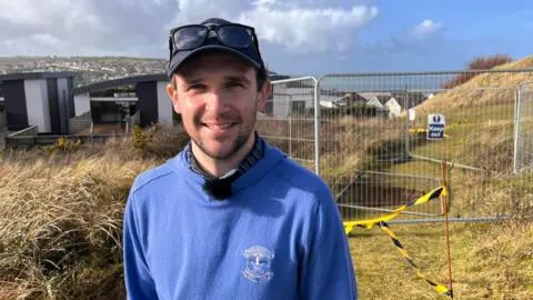 Man in a blue woollen jumper, wearing a cap with sunglasses resting on top, in front of a fenced off mine hole in a grassy area