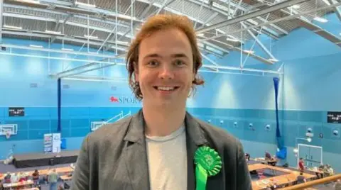 Peter Charles Rees smiles at the camera as he stands in an elevated position in a sport hall, with people sitting at tables during the local election count.