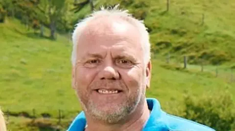 Mark Williams stands in a field. Mark has close-cropped grey hair and a short beard and he's smiling into the camera. He is wearing black and blue T-shirt.