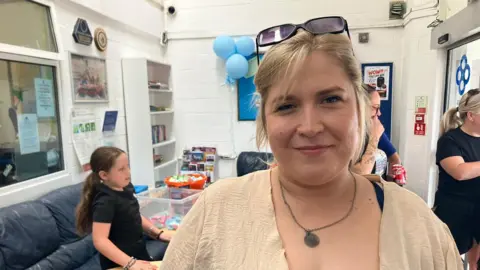 Lucy Regan stands in a room, she is wearing a light brown top over a denim dress and is wearing a necklace