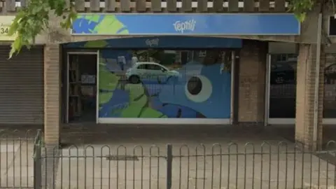 Google A shop on a precinct with a blue banner across it and the words reptili in white writing. It is within a brown brick building and there is a black fence surrounding the area.