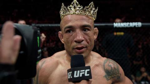 Jose Aldo wears a crown as he speaks on a UFC microphone after a fight