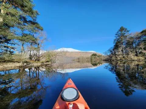 Doug Kinnaird The front of an orange kayak on a very calm Loch Awe. There is a snow-capped mountain in the background reflected in the water which is bright blue, There are trees on either side, also reflected in the water.