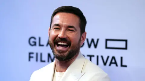 PA Media Martin Compston laughing while on the red carpet at the Glasgow Film Festival - a banner with the festival name is behind him, and he is dressed in a white suit