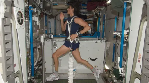 Wanita NASA dengan celana pendek Angkatan Laut dan teratas di atas treadmill yang diikatnya melalui harness bungee putih.