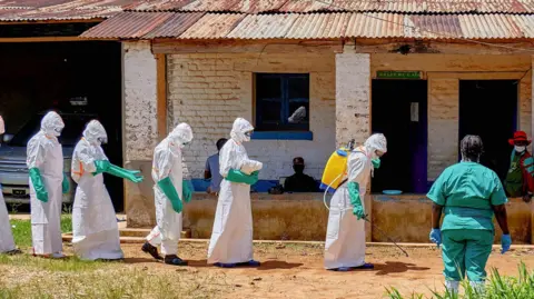 Five Red Cross workers in full PPE - white suits and green rubber gloves - walking in a straight line while the one in front is carrying a canister on their back and spraying disinfectant on the ground. They are outside Mongwalu General Hospital and two people are watching them.