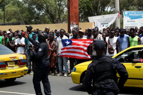 Abdul Baha Jallanku / EPA men waving the flag of Liberia in the police, who stand across the road. In the middle there are yellow taxis.