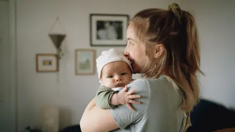 A woman faces to the left of the picture, holding a baby, who is looking at the camera 