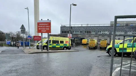 Ambulances parked in a car park outside of a hospital. The hospital building is large and grey. A large red sign is displayed in front of the car park which reads 'emergency vehicles only beyond this point'. 