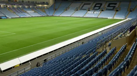 Blue players seats next to the player's entrance, seen from pitch side at a football stadium. Beyond, premier and hospitality zones can be seen.