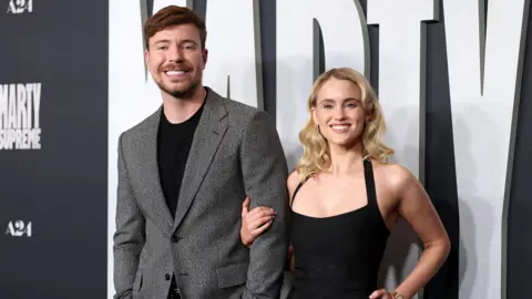 Getty Images MrBeast, real name Jimmy Donaldson, and his partner Thea at an event. He is wearing a grey suit jacket over a black tshirt. She is wearing a black strappy dress. 