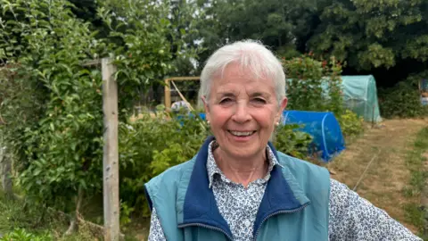 A woman with white hair and a green and blue gilet, wearing a flowery shirt underneath. She is smiling at the camera. In the background, you can see her allotment.