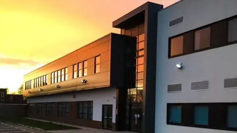 Image shows the outside of Melton Theatre which is a white and black modern looking building. The sun is setting over the building which is reflecting on the windows and giving it a warm yellow, orange glow. 