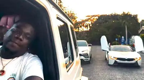 ABUTRICA/INSTAGRAM Abu Trica in a white Mercedes G-wagon, near other luxury cars.