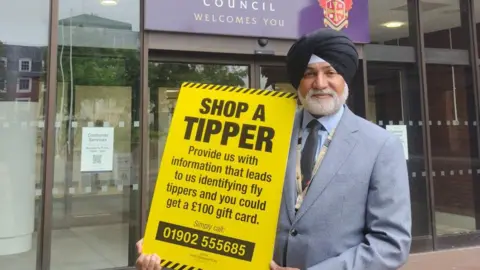 A man in a grey suit and a dark turban smiles at the camera while holding a yellow sign reading: "Shop a tipper. Provide us with information that leads to us identifying fly tippers and you could get a £100 gift card."
