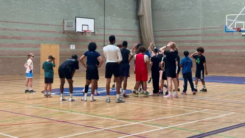 NSVPP A large group of people, standing in a sports hall, all with their backs to the camera, looking to be playing basketball. There are hoops on the wall and a net in the corner. 