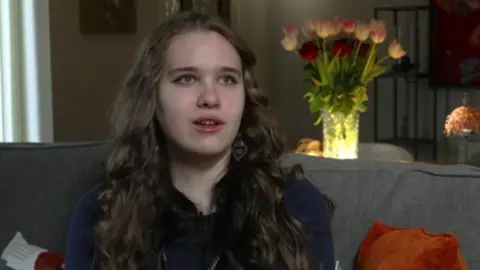 A young woman sits on a grey sofa with an orange cushion on her left. She has long, curly brown hair. A vase filled with pink tulips and red roses can be seen on a table behind the sofa.