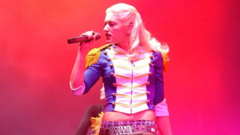 Gwen Stefani sings, she has long blonde hair and is wearing a white, blue and gold jacket.