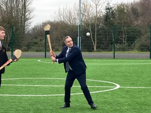 BBC Paul Givan playing hurling with a pupil on a sports field. Both are holding hurling sticks - Givan is tossing a sliotar in the air and preparing to hit it. He is wearing a dark blue suit, light blue shirt and sliver tie.