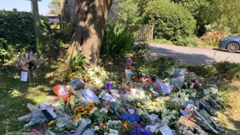 Dozens of bouquets of flowers laid on the grass in front of a tree near a hotel. Among the floral tributes at the back is a black and white photo of Widdecombe.