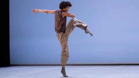 Gregory Batardon & Rodrigo Buas Jakob dancing, on a stage, with a blue background. He is wearing a brown top, brown trousers and grey socks. His left arm is outstretched and right arm across his body. His leg is raised. He has short curly dark hair. 