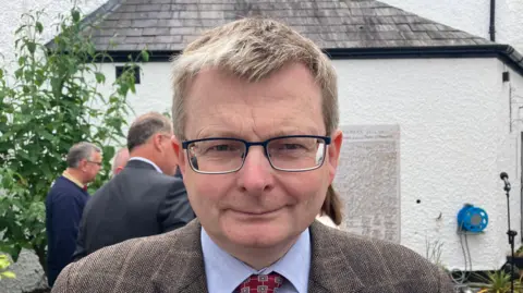 Dr William Roulston is wearing a black-rimmed glasses and is wearing a tweed suit. He has a blue shirt and red tie.