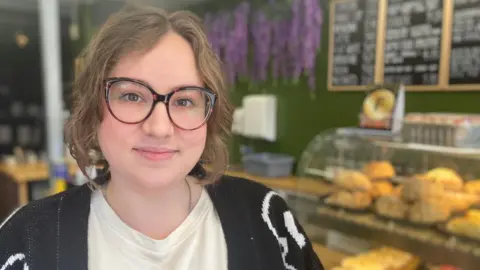 A head and shoulders shot of a woman with short brown hair and large tortoiseshell glasses. She is wearing a white t shirt and black cardigan and standing in front of a bakery counter.