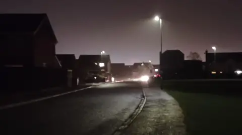 A very dark street with a street light ahead on the left hand side. The right hand side of the road is dark.