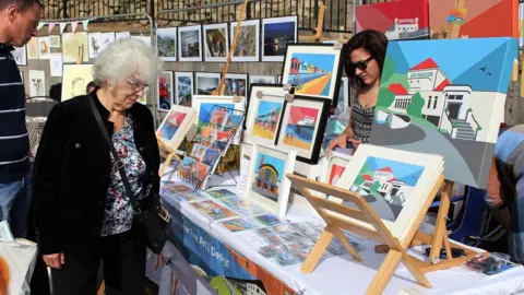A woman browses a stall an an open-air art market. A woman with black hair and sunglasses stands behind the stall. 