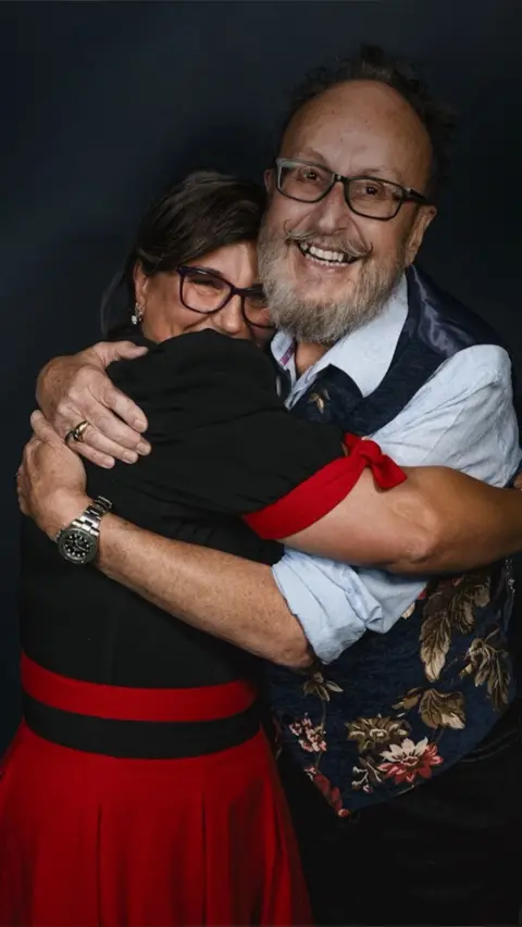 Lili Myers and Dave Myers stand closely together in a warm embrace, both smiling. Lili wears glasses, a black top with red accents, and a red skirt. Dave also wears glasses along with a light blue shirt and a dark floral-patterned vest. The background is dark, drawing focus to their affectionate pose and expressions.