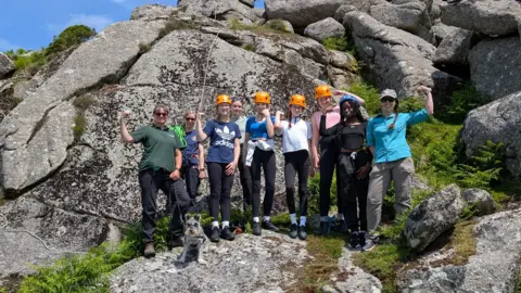 BBC The group of women and girls who took part in the activity all stand against the rockface with their arms raised in Rosie the Riveter pose.