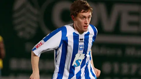 Paul Hazlewood/Bhafc Jake menggiring bola dengan bola di Withdean, stadion Brighton saat itu, pada tahun 2009. Dia mengenakan kit rumah biru dan putih klub yang memiliki kaus kaki dan celana pendek putih. Bola berwarna biru, merah dan putih. 