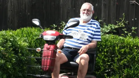 Bob is wearing a blue-and-white hooped top over dark blue shorts. He has white hair and a beard, and is sitting on his mobility scooter, which is red with a black wire basket on the front. He is in his garden with a green hedge behind him.