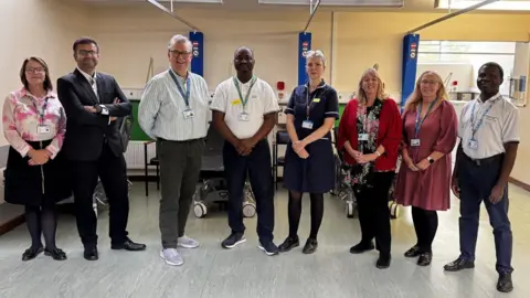 Mid and South Essex NHS A group of four men and four women standing in a line on a hospital ward. They are all smiling.