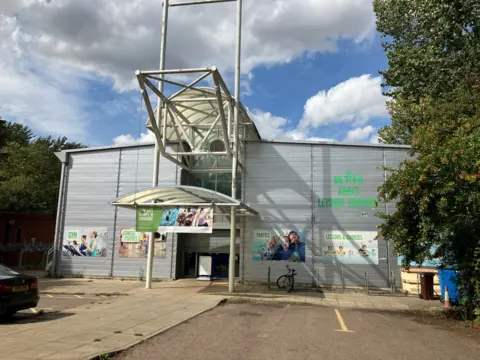 Exterior of Abbey Leisure Complex. The building is a silver-coloured warehouse-type building. Cars are parked and there's a bicycle outside. A woman is leaving the building.