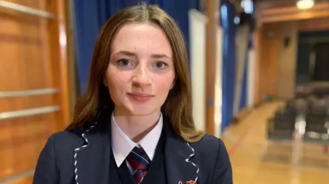 Francesca,  who is wearing a navy school uniform, has light long brown hair, and dark brown eyes, looks at the camera with a neutral expression