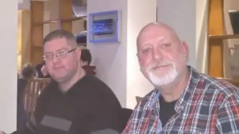 Sarah Merritt Two men - one wearing a dark jumper and the other wearing a chequered shirt - sitting next to each other in what looks like a restaurant. They are looking into the camera.