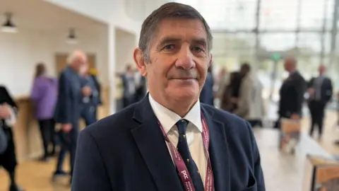 BBC Philip Wilkinson looks into camera wearing a suit and tie in a room with others