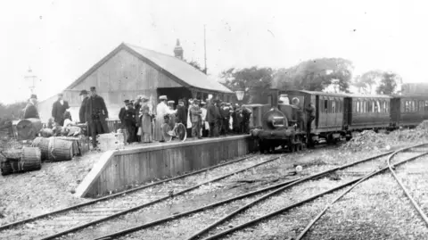 Manhood Wildlife & Heritage Group A historic photograph of a station on the Hundred of Manhood and Selsey Tramway. Passengers wait on the platform beside a steam train, while luggage and parcels are stacked nearby. Several railway tracks are visible in the foreground.
