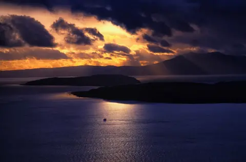 Douglas Soutter The outlines of two islands are just about visible in a striking sunset with orange-yellow light piercing through dark clouds