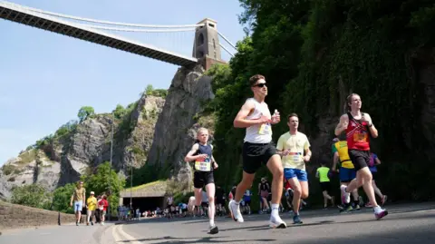 People in running gear are running on a road. In the background we can see the Clifton Suspension Bridge.