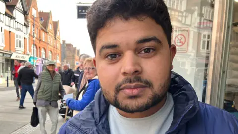 Vikki Irwin/BBC A man wearing a blue jacket. He is on a street with people behind him, and he is looking at the camera.
