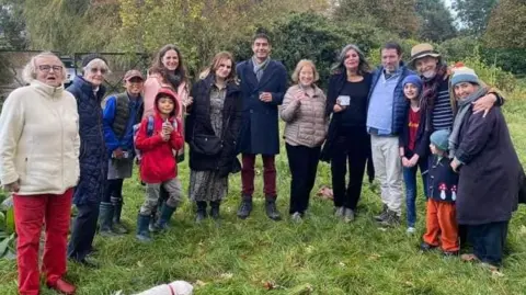 A group of 14 smiling people on the site of Park Road Allotments. They are of varying ages and a dog is running around the background.