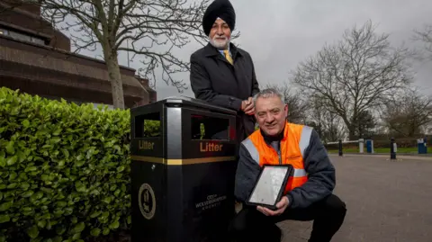 Wolverhampton City Council Councillor Bhupinder Gakhal, cabinet member for resident services at City of Wolverhampton Council, with Andy Moore, public realm team leader. They are by a litter bin in the city with Mr Moore holding some of the technology linked to the sensors. Mr Gakhal is leaning on the bin with Mr Moore crouched beside it.