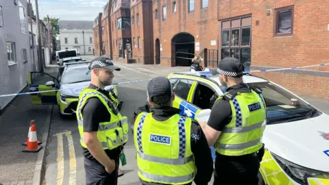 Three police officers standing beside a police car on Bedford Street. There is a police cordon in place and the road is closed.
